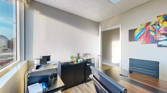 Office 201 with a wood-tone desk, black chairs, a mini fridge, glassware and colorful lion artwork.