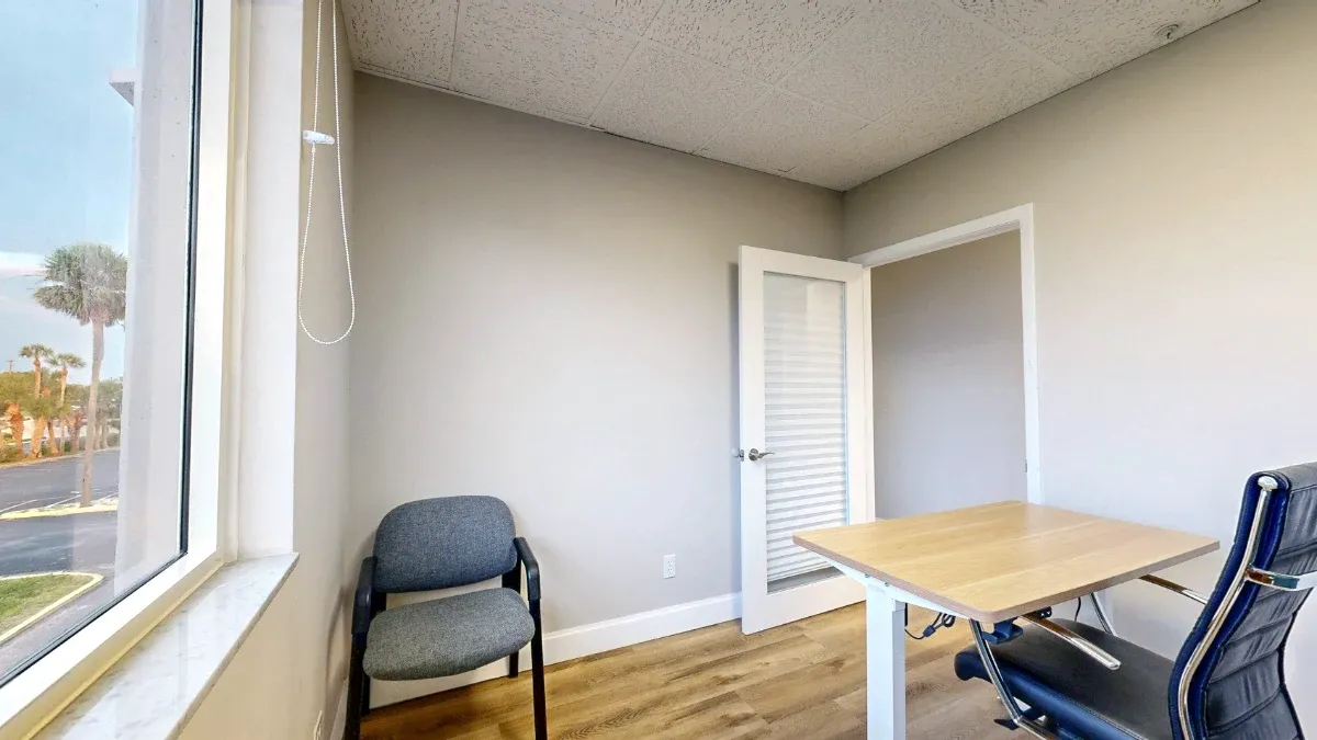 View from the window side of office 204 shows a gray guest chair, wood-top desk, black office chair and open door.