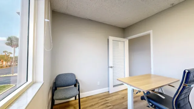 View from the window side of office 204 shows a gray guest chair, wood-top desk, black office chair and open door.