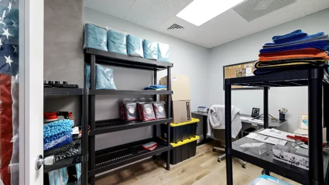 Inside Office 217, storage shelves and folded cloths frame a rear desk with a rolling chair and stacked bins.