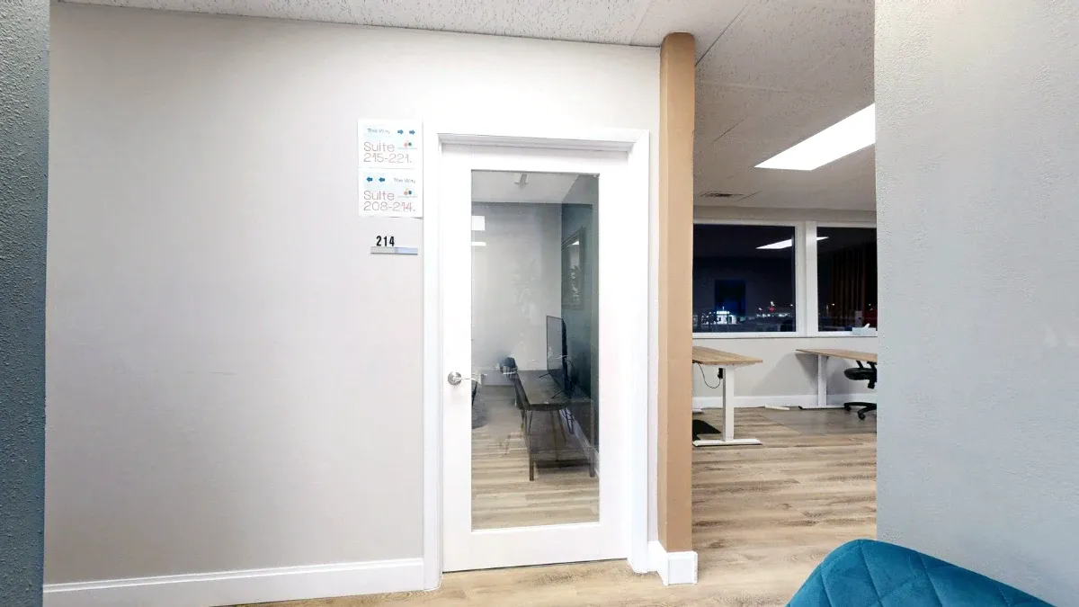 White glass-paneled door marked 214 beside directional signs, with desks visible in the adjoining workspace.