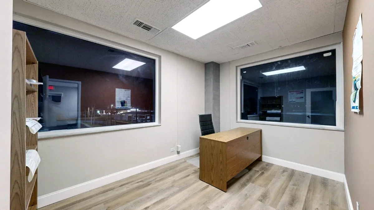 Office 216 interior with two large windows, a wooden desk, a black chair and open floor space.