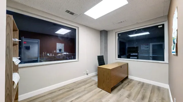 Office 216 interior with two large windows, a wooden desk, a black chair and open floor space.