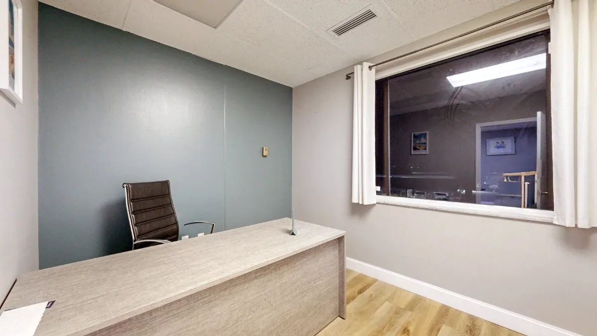 Private office with a light wood desk, black chair, blue accent wall, and large interior window framed by white curtains.