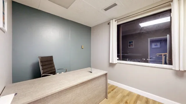 Private office with a light wood desk, black chair, blue accent wall, and large interior window framed by white curtains.