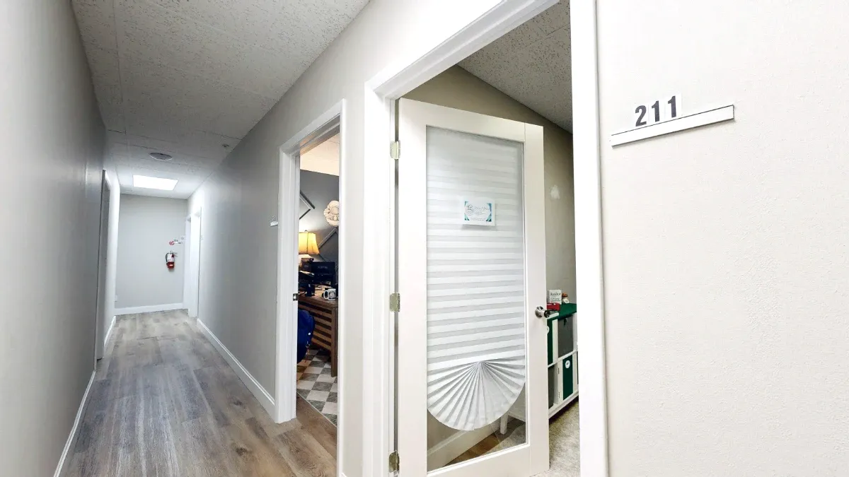 Hallway view of Office 211 with its number plaque and open glass-panel door fitted with a pleated shade.