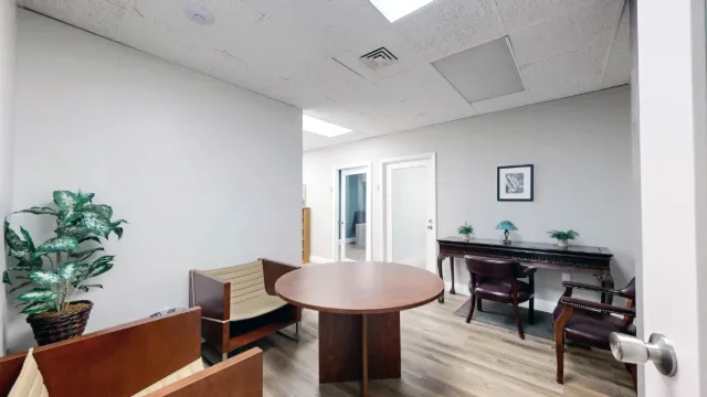 Doorway view of a round wooden table, cushioned seating, a tall plant, and a console desk in the Suite 222 shared area.