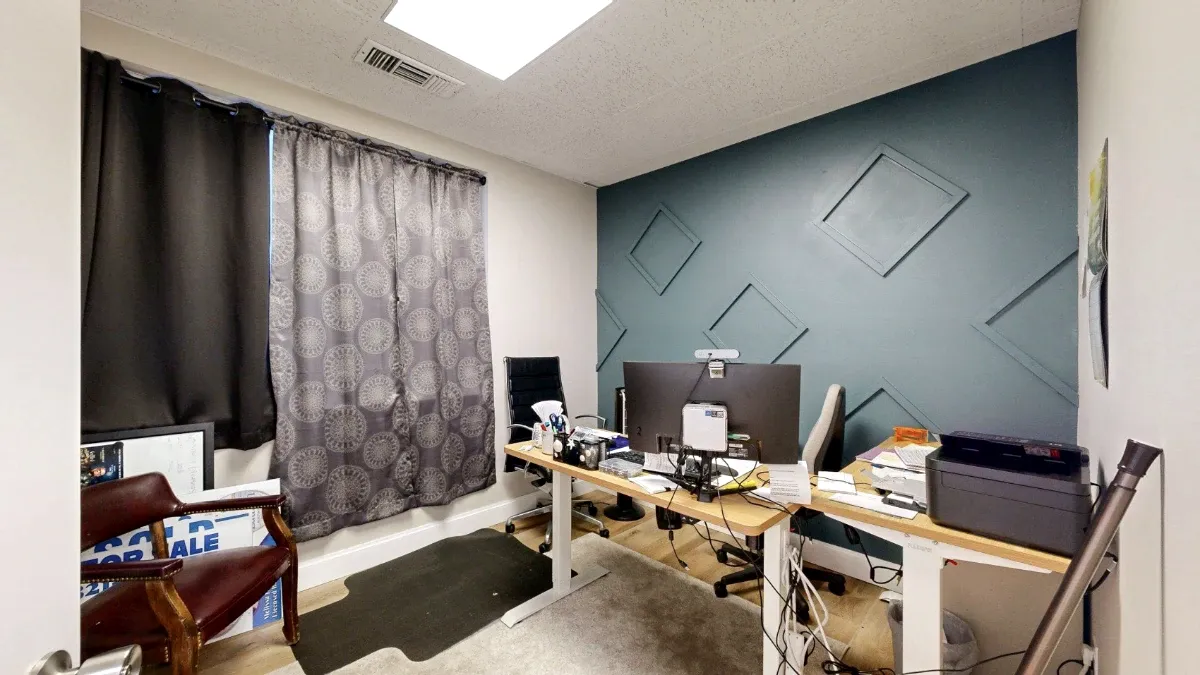 Office 209 viewed from the doorway, with a curtained window, desk, printer, guest chair and blue geometric accent wall.