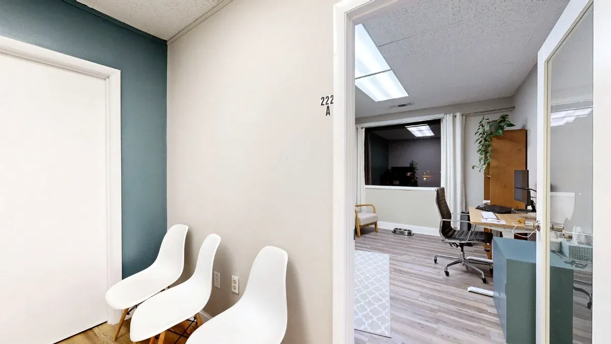 Three white hallway chairs beside the open door to Office 222A, with a desk, black office chair and window visible inside.