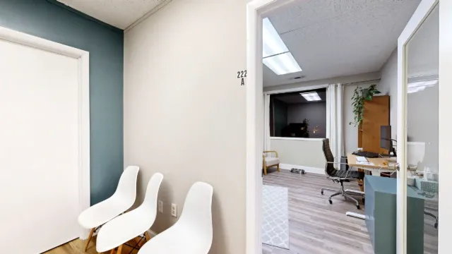 Three white hallway chairs beside the open door to Office 222A, with a desk, black office chair and window visible inside.