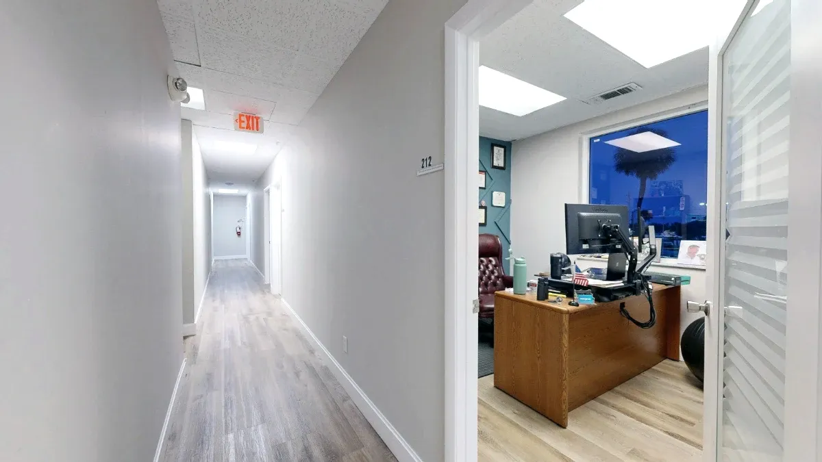Hallway view into Office 212, showing the numbered doorway, wooden desk, monitor and large window.
