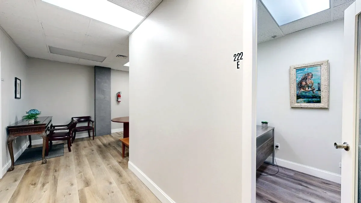 Shared hallway seating beside office E, where a desk, small plant, and framed coastal artwork are visible.