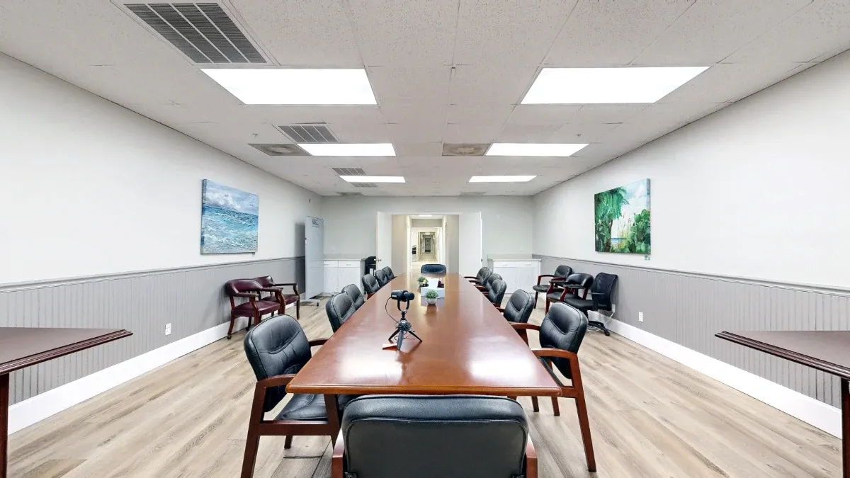 View down the wooden conference table toward the hallway, with a tabletop tripod, padded chairs and coastal paintings.