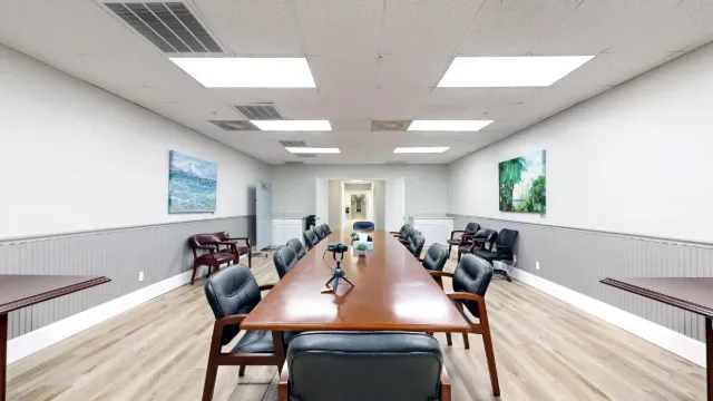 View down the wooden conference table toward the hallway, with a tabletop tripod, padded chairs and coastal paintings.