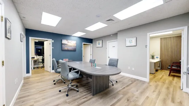 A rounded conference table with gray rolling chairs sits among office doorways beneath a blue accent wall and coastal art.