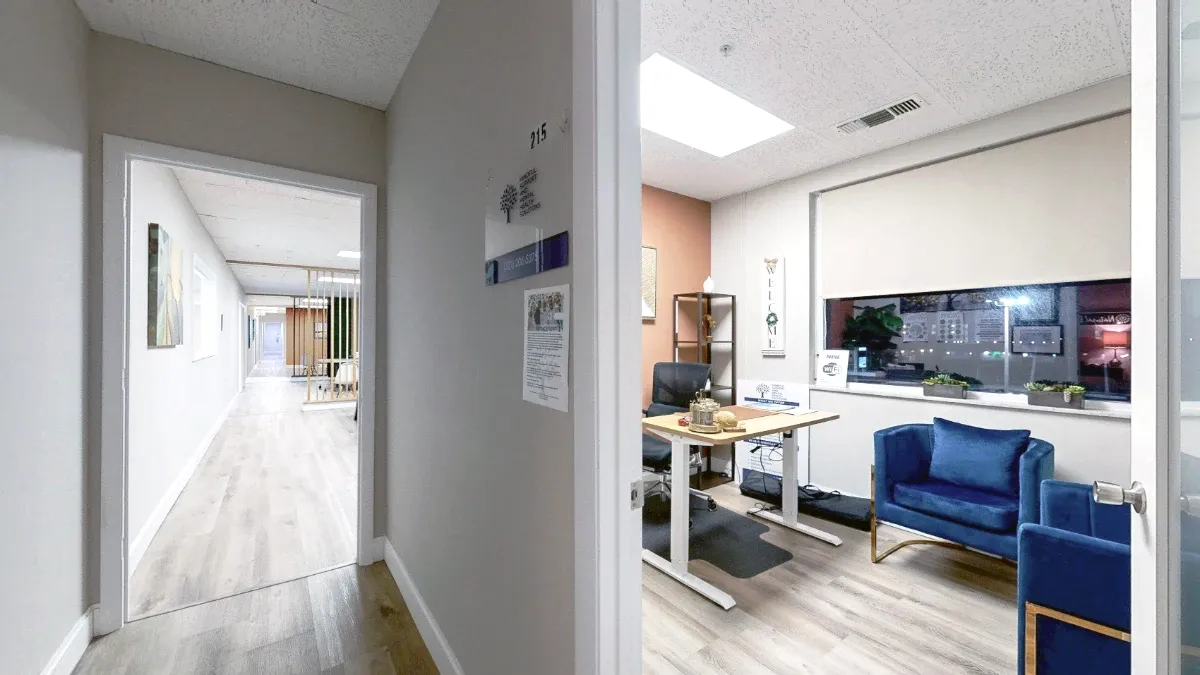 Angled entrance view of Office 215 showing a desk, blue seating and a wide window with a partially lowered shade.