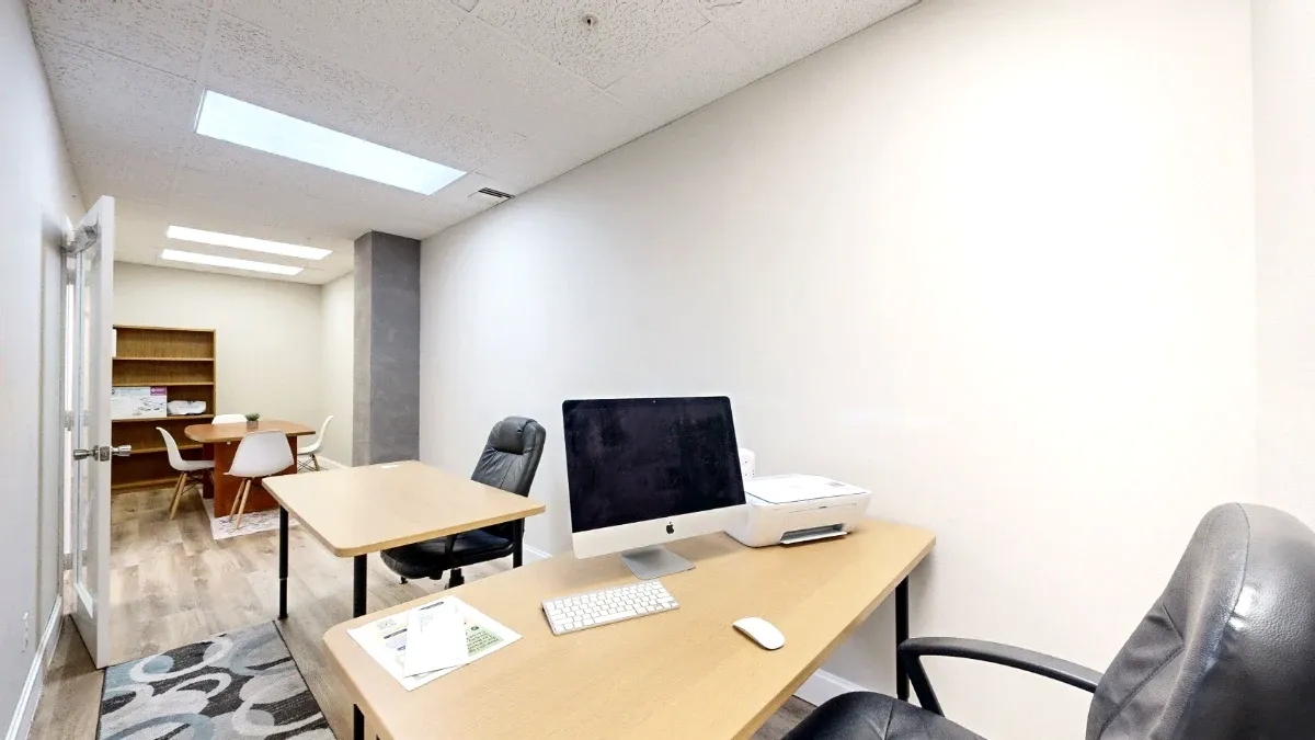A computer and printer sit on the foreground desk, with a second desk and a table with white chairs farther back.