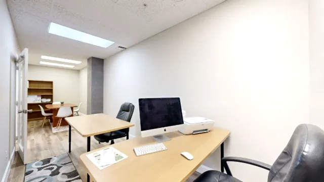 A computer and printer sit on the foreground desk, with a second desk and a table with white chairs farther back.