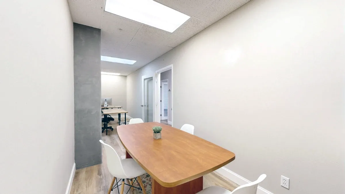 A wooden table with white chairs and a small plant fills the foreground, with a gray column and desks beyond.