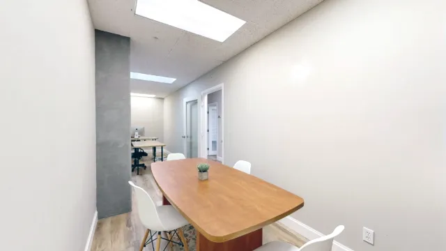 A wooden table with white chairs and a small plant fills the foreground, with a gray column and desks beyond.