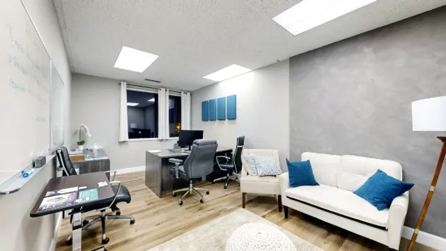 Wide view of Office 224A showing a desk beneath curtained windows, office chairs, a whiteboard and a sofa.