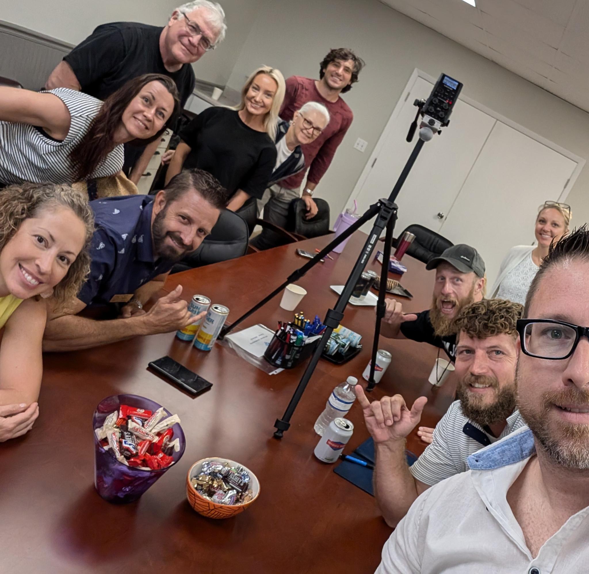 A group smiles around a conference table with a tripod-mounted recorder, drinks, candy bowls and office supplies.