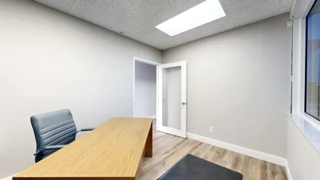 Small office with a wood desk, gray task chair, window, wood-look flooring, and an open glass-paneled door.