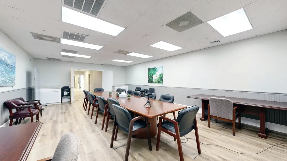 Diagonal view across the meeting table toward the open entrance, with side tables and extra chairs along the walls.