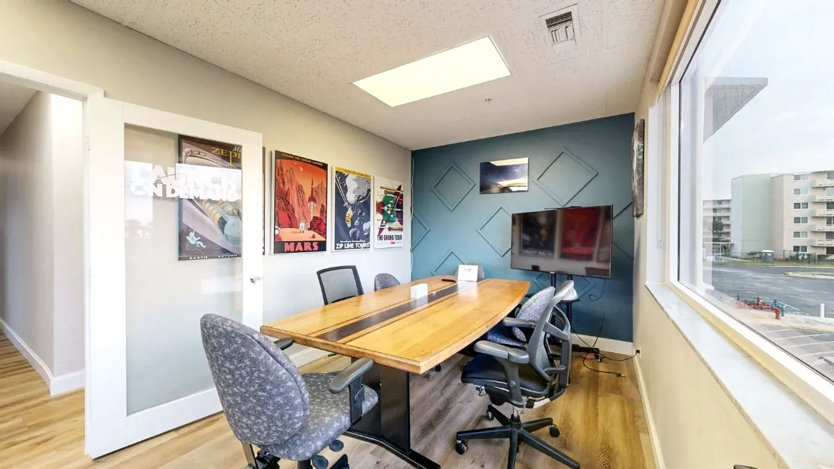 Wooden meeting table surrounded by rolling chairs in office 203, with a screen, teal accent wall, and large window.