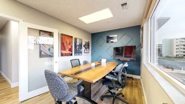 Wooden meeting table surrounded by rolling chairs in office 203, with a screen, teal accent wall, and large window.