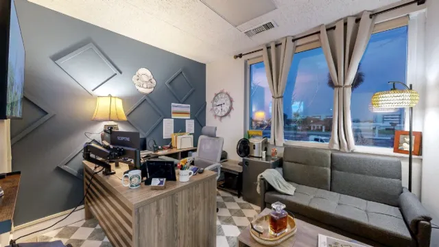 Wood-finish desk and gray chair beside a sofa beneath curtained windows, with geometric trim on the accent wall.