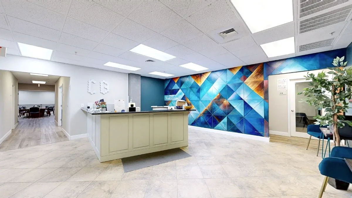 Wide reception view with a central white desk, geometric mural, left hallway, and blue seating beside a leafy plant.