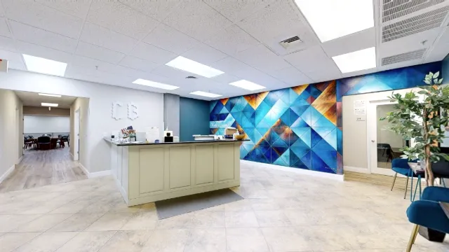 Wide reception view with a central white desk, geometric mural, left hallway, and blue seating beside a leafy plant.