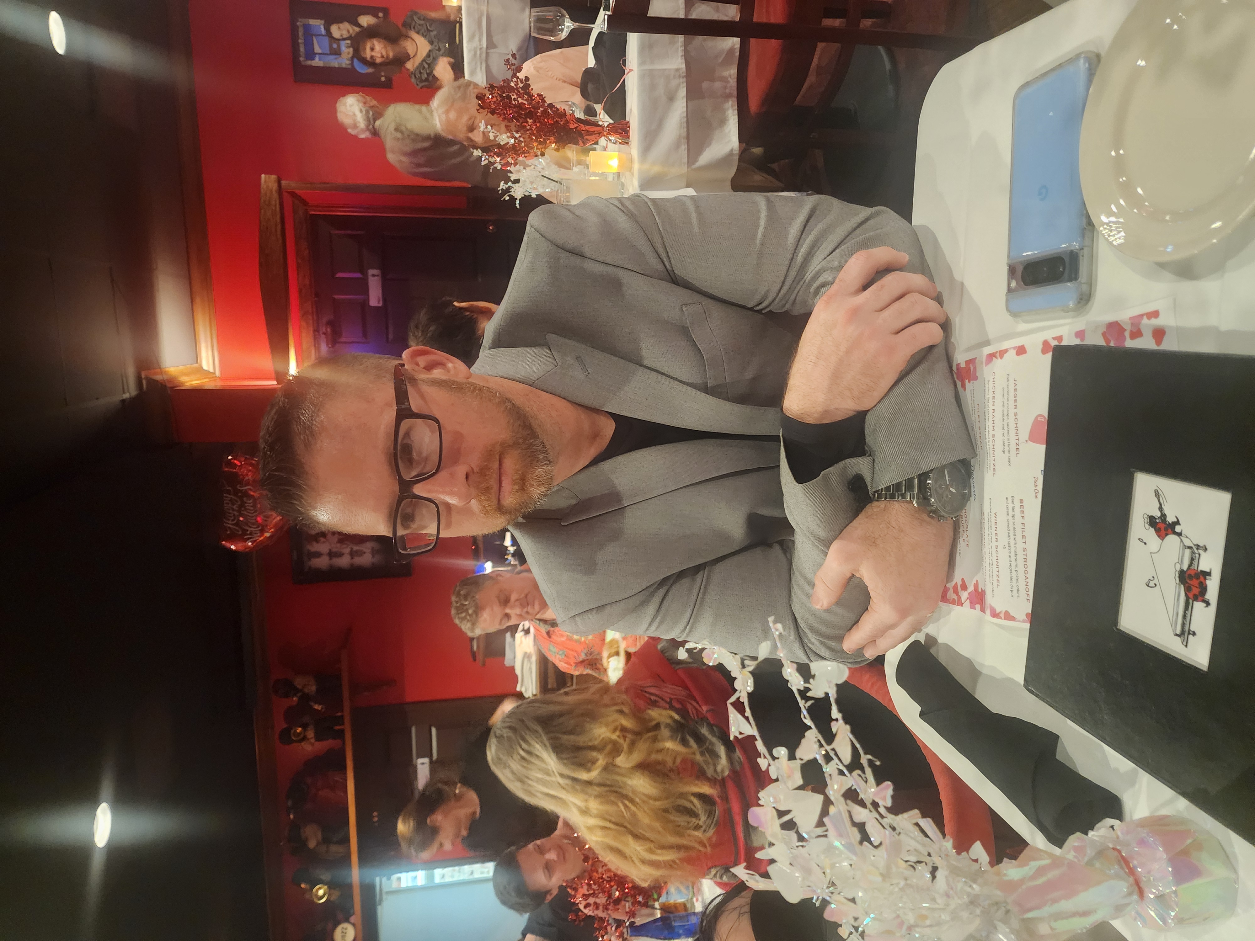 A man in glasses and a gray blazer sits at a restaurant table surrounded by diners and Valentine decorations.