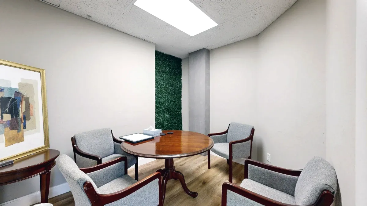 Four gray armchairs surround a round wooden table in The Cape, with framed artwork and a green accent panel behind.