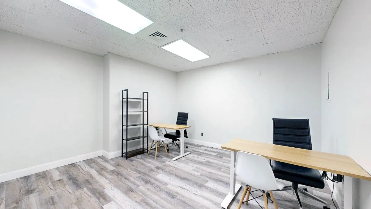 Two wood-top desks with black office chairs and white guest chairs flank an open floor area, with black shelving at the back.