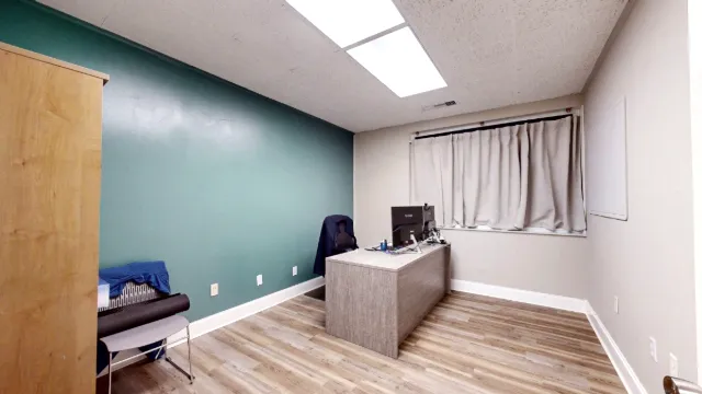 Private office with a teal accent wall, desk, monitors, chairs, wood-look flooring and a curtained window.