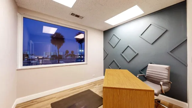 Office with a wood desk, gray chair, geometric accent wall, and a large window overlooking A1A and palm trees.