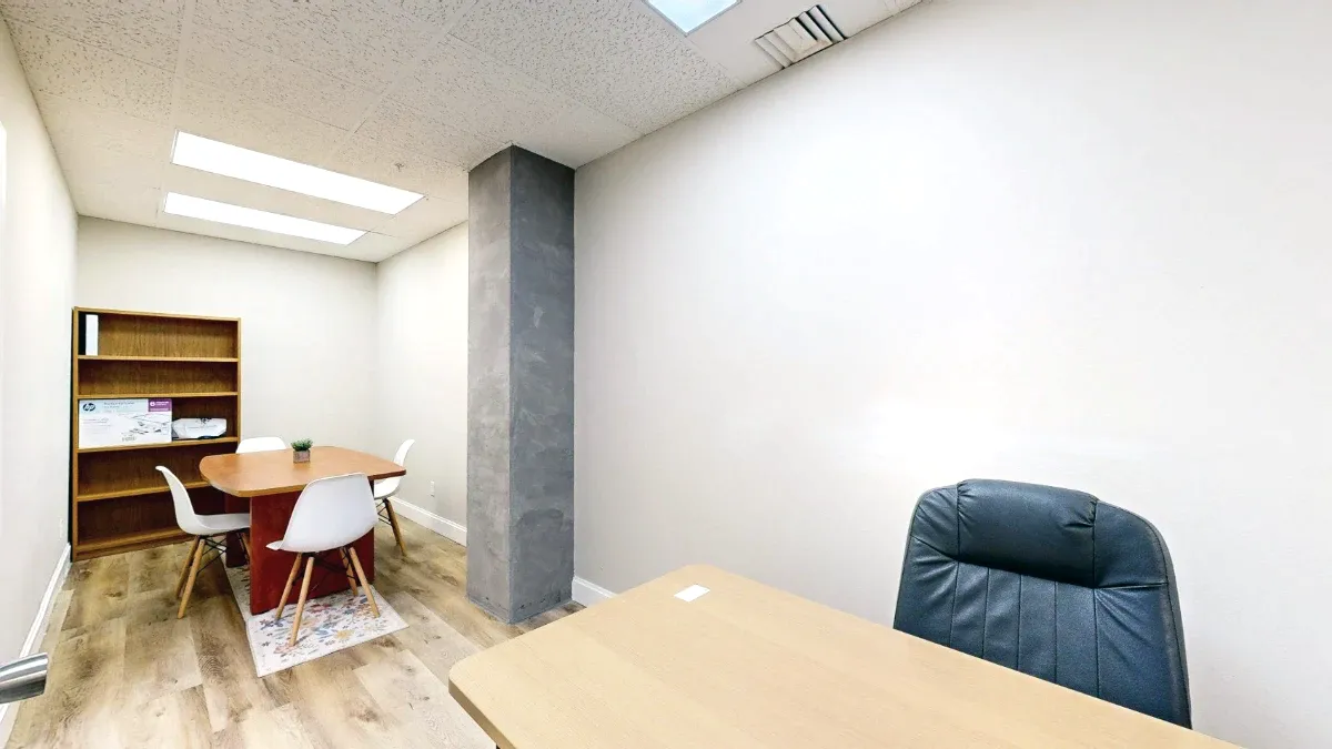 A desk and black chair face a rear table with white chairs, a wooden bookcase and a tall gray column.