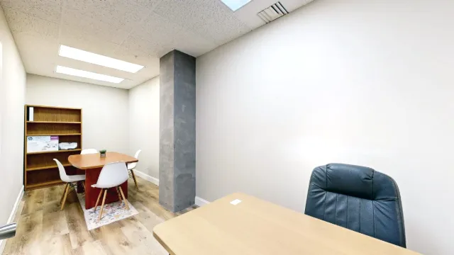A desk and black chair face a rear table with white chairs, a wooden bookcase and a tall gray column.