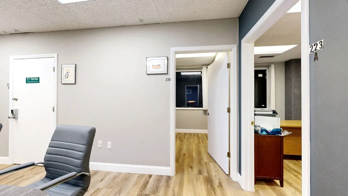 A gray chair fills the foreground beside open doorways marked 220 and 223A, with office furniture visible beyond.