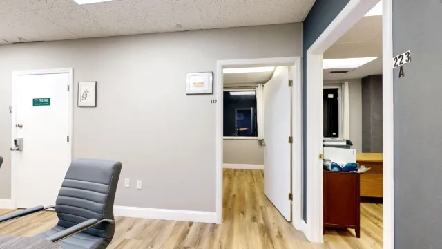 A gray chair fills the foreground beside open doorways marked 220 and 223A, with office furniture visible beyond.
