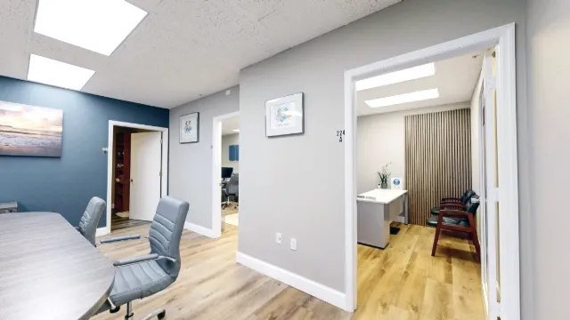 Gray conference chairs fill the foreground beside an open office with a white-topped desk, dark guest chairs, and slat wall.