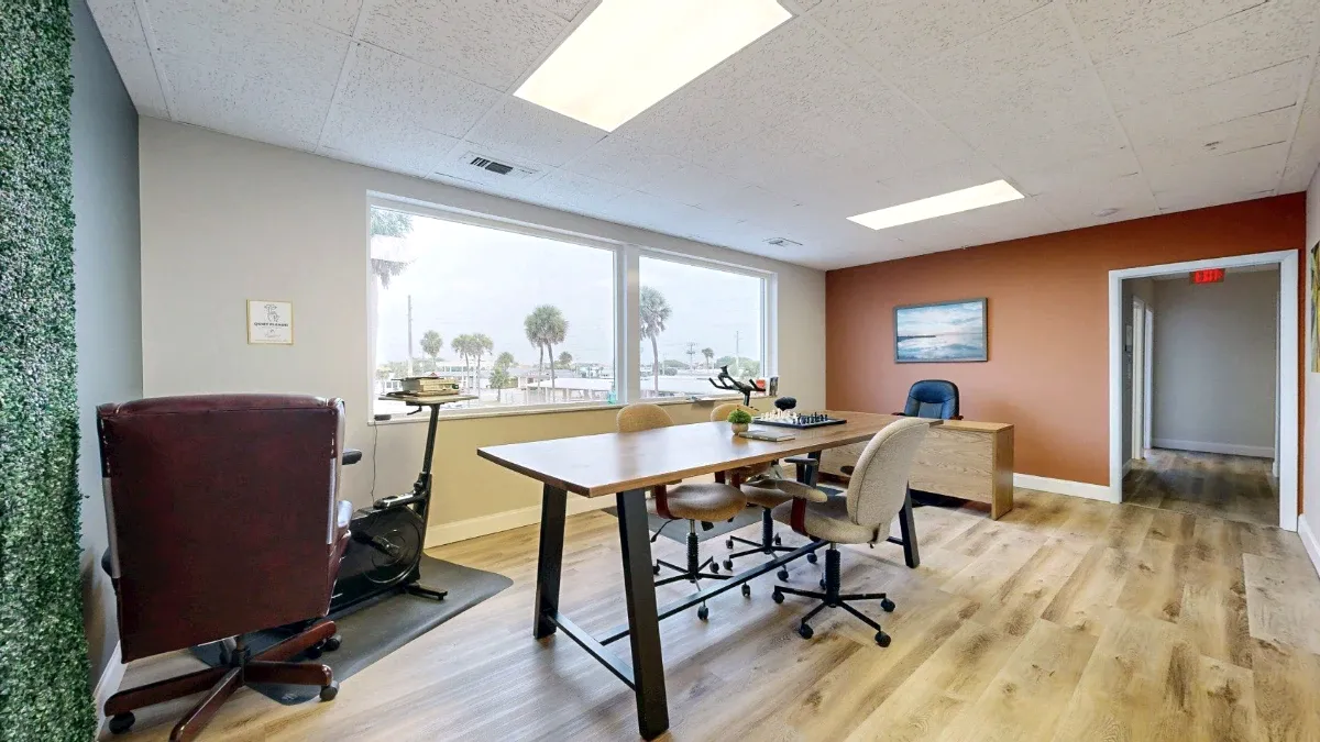 A tall shared table with a chessboard sits between window-side bike seating and a desk against an orange wall.